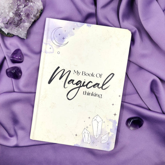 "My Book Of Magical thinking" muistivihko violetilla taustalla ametistiryväksen ja rumpuhiottujen ametistien kanssa